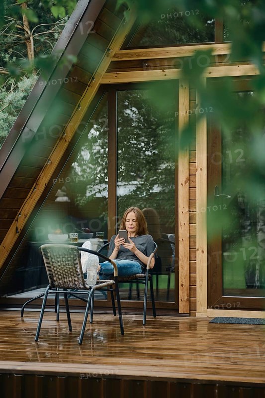 Serene moment after the rain. Pretty woman is reading something on smartphone. She is seated comfortably outside a modern wooden cabin with large glass windows, surrounded by lush greenery