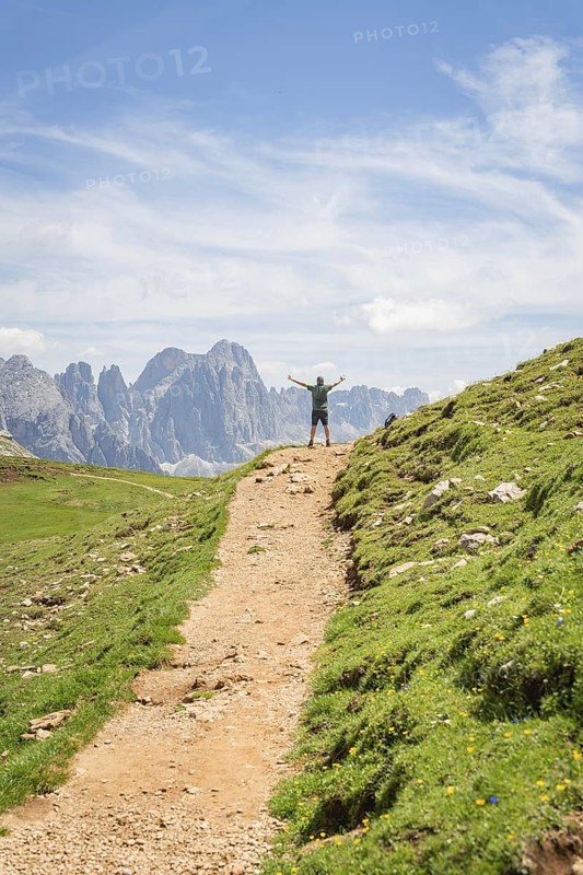 A hiker on a mountain path with outstretched arms in front of an impressive mountain scenery, Schlehrn hike, Alpe di Siusi, Dolomites, South Tyrol, Italy