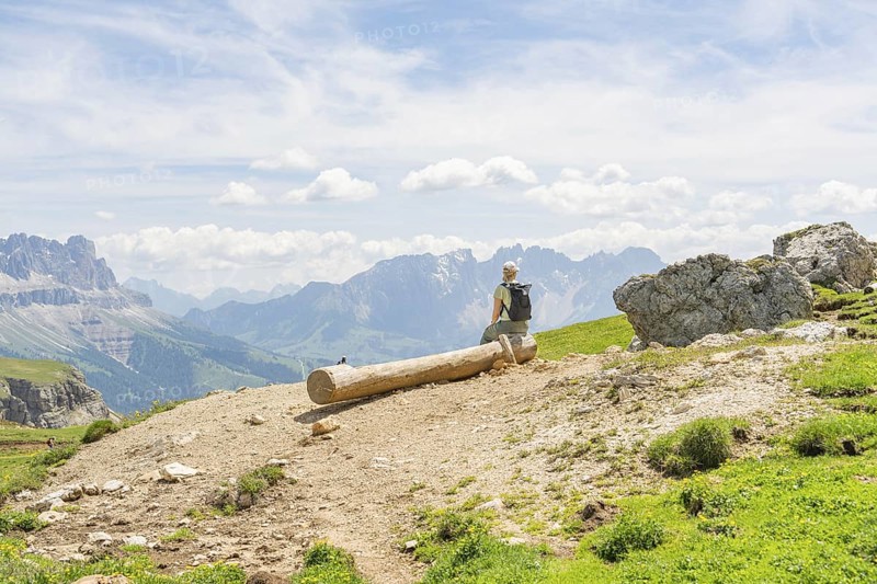 Hiker sitting on a tree trunk with a wide view over the mountains and valleys, Schlehrn hike, Alpe di Siusi, Dolomites, South Tyrol, Italy