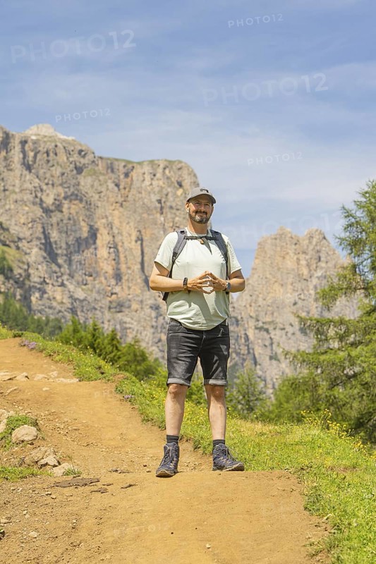 A man with a rucksack stands smiling on a mountain path with a rocky background, Schlehrn hike, Alpe di Siusi, Dolomites, South Tyrol, Italy