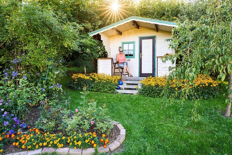A woman sits relaxed on the small veranda of a garden shed and reads. Blooming summer flowers in the garden. Germany