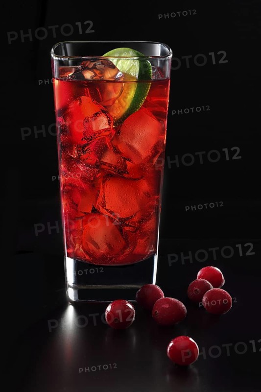 Cocktail in a long drink glass on ice with a slice of lime and cranberries as garnish against a black background, studio shot