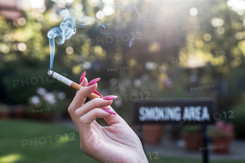 Woman's hand with long pink nails holding a cigarette. Blurred smoking area sign in background. Generative ai, AI generated