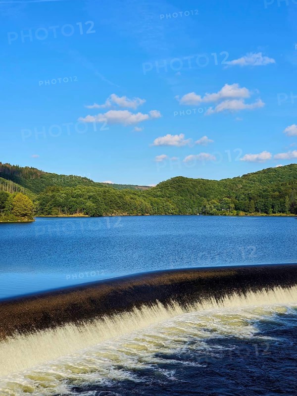 A quiet reservoir with a small waterfall, surrounded by wooded hills under a blue sky, summer, Rursee, Obersee, Rurberg, Simmerath, Eifel National Park, North Rhine-Westphalia, Germany