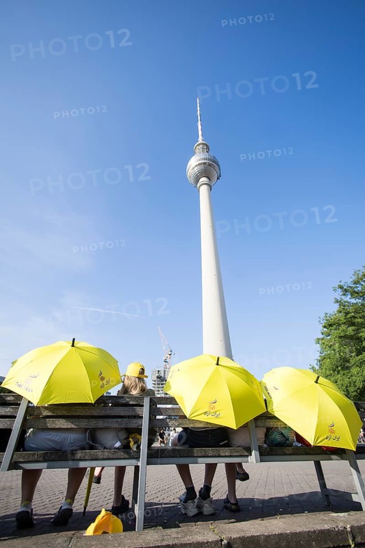 Several people sit with yellow Free Iran umbrellas in front of the television tower on Alexanderplatz in Berlin on 21 June 2025