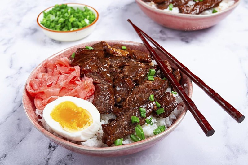Gyudon Japanese beef with rice, pickled ginger, boiled egg slice, garnished with green onions, in a bowl with chopsticks