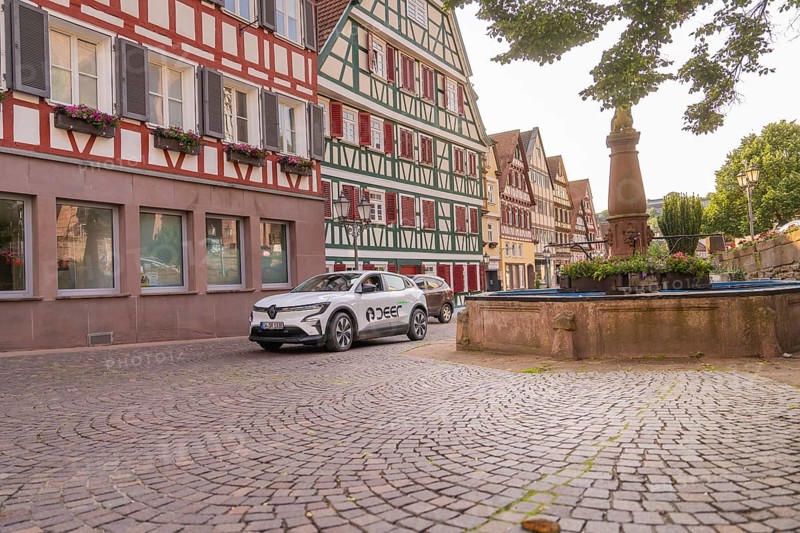 A car next to a fountain in a cobbled old town street with half-timbered facades and green plants, Deer E, car sharing, electric car, Calw, Black Forest