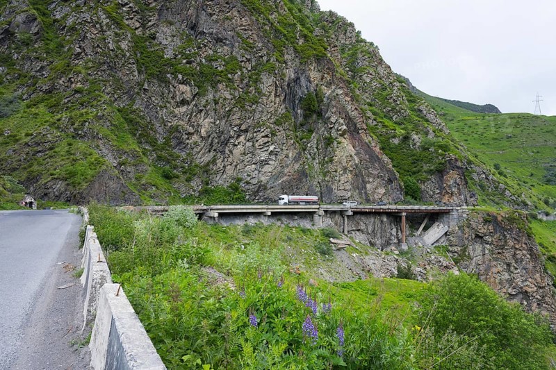 A truck crosses a bridge along steep rock faces in a green mountain landscape, damaged concrete road patched with iron pipes, Kazbegi, Stepantsminda, Georgian Military Highway, Mtskheta-Mtianeti Province, High Caucasus, Georgia