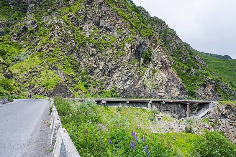 A road runs along steep rock faces, with a bridge and green vegetation, damaged concrete road patched with iron pipes, Kazbegi, Stepantsminda, Georgian Military Highway, Mtskheta-Mtianeti Province, High Caucasus, Georgia
