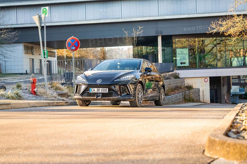 Black car drives in front of a modern building backdrop during the day, road signs show the way, MG4 electric car, deer e-car sharing, Calw, Germany