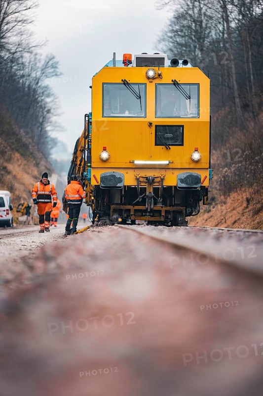 Close-up of a yellow train with workers on rails under cloudy sky, using a tamping machine on the Hermann Hessebahn construction site, Calw, Germany