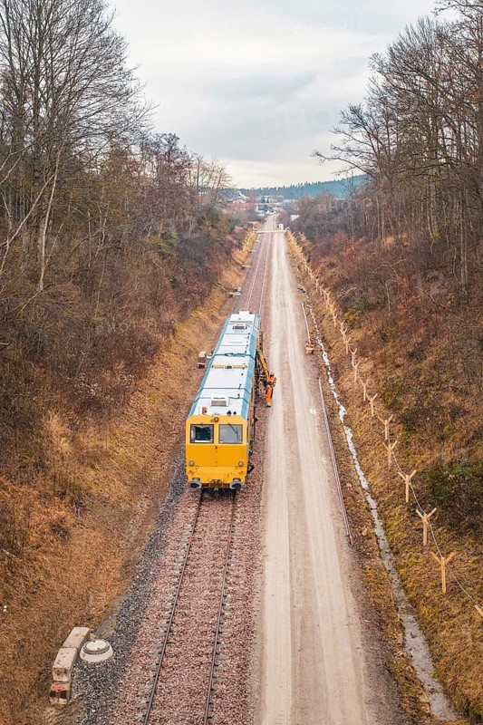 Yellow train during construction on a long rail line surrounded by bare trees, using a tamping machine at the Hermann Hessebahn construction site, Calw, Germany