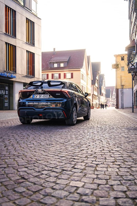 Black car drives through a paved city street between modern and historic buildings in sunlight, MG4 electric car, Deer e-Carsharing, Calw, Germany