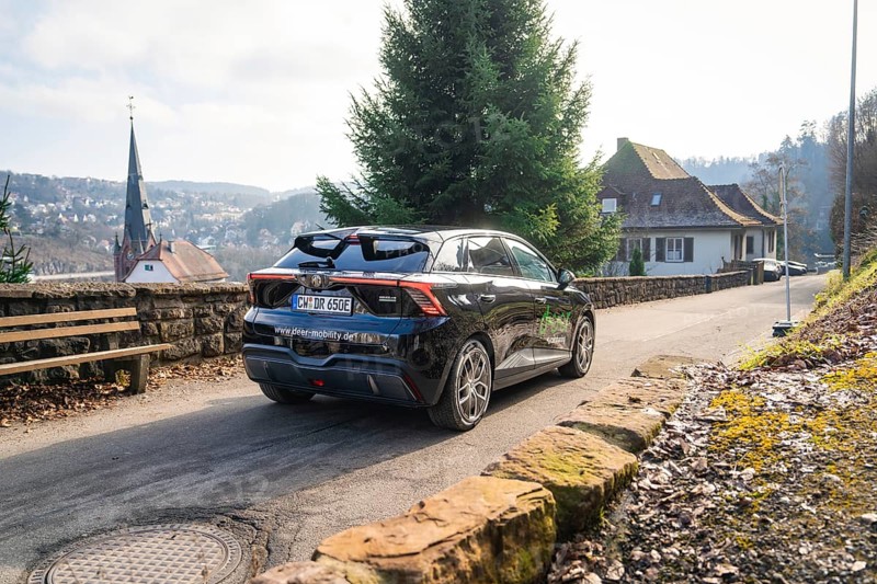 Car driving on a rural road with house and hills in the background, MG4 electric car, Deer e-Carsharing, Calw, Germany