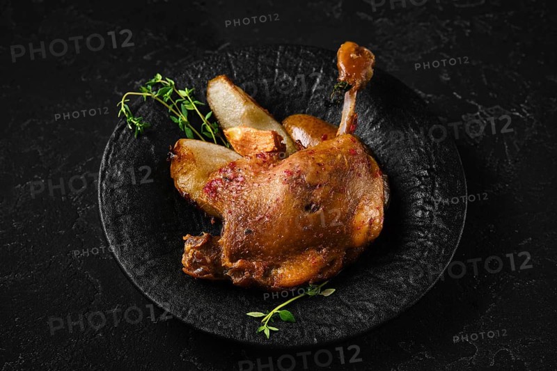 Duck confit on a black stone plate with pieces of roasted pear and herbs. The setting shows a dark background. The dish highlights the slow cooking method and texture of the meat