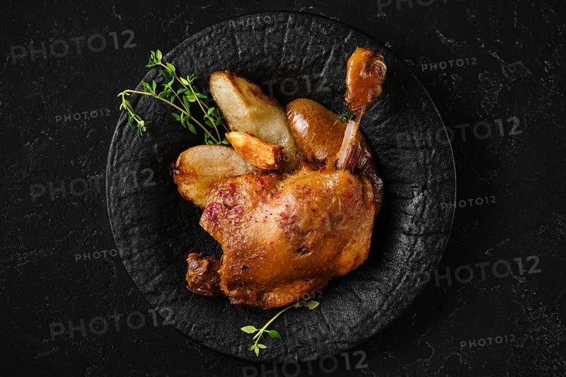 Top view of roasted duck leg on a textured black plate with caramelised in duck fat slices of pear. The setting is a simple kitchen space with a dark background, highlighting the meal's details