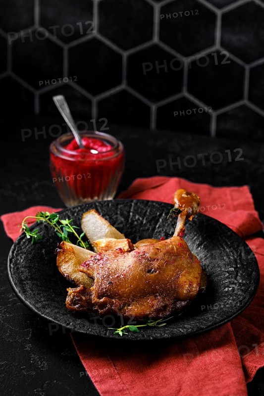 Roasted duck leg with crisp skin and tender meat on a stone plate. A small jar filled with a lingonberry sauce sits nearby. Green herbs are placed on the side of the duck on a dark table