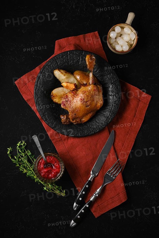 A plate holds a portion of duck confit alongside roasted pears. A small bowl of lingonberry sauce and pickled pearl onions on the table. Fresh herbs are placed next to the plate