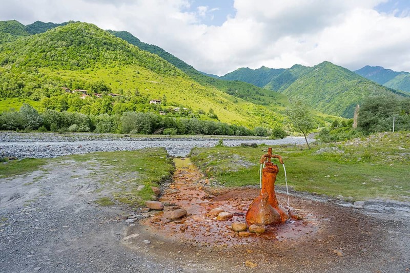 A rusty spring flows onto a gravel riverbed surrounded by green mountains, mineral spring near Nadibani, White Aragvi River, Mtskheta-Mtianeti region, Georgian Military Highway, High Caucasus, Georgia