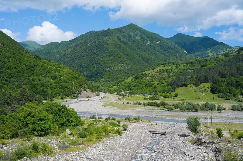 A riverbed leads through green mountains under a partly cloudy, blue sky, landscape near Chartali, White Aragvi River, Mtskheta-Mtianeti region, Georgian Military Highway, High Caucasus, Georgia