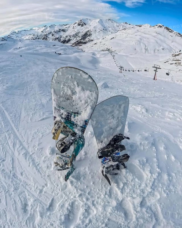 Snowboards rest in deep snow on a high mountain summit, offering a panoramic view of an expansive ski resort with distant peaks, snowy slopes, and a chairlift system under a bright winter sky