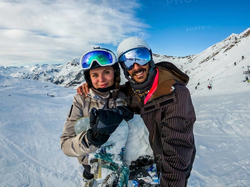Smiling young couple in winter sportswear and goggles embracing with a snowboard, standing on a snowy mountain slope with ski lifts and distant peaks under a clear blue sky
