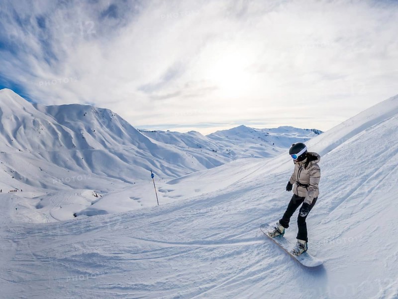 Female snowboarder in helmet and goggles carving fresh snow on a sunny alpine slope, panoramic mountain vista behind her conveying adventure, speed and open sky freedom