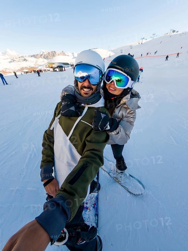 Couple having fun snowboarding on a sunny winter day, enjoying their active vacation in the snow covered mountains while capturing a joyful selfie together