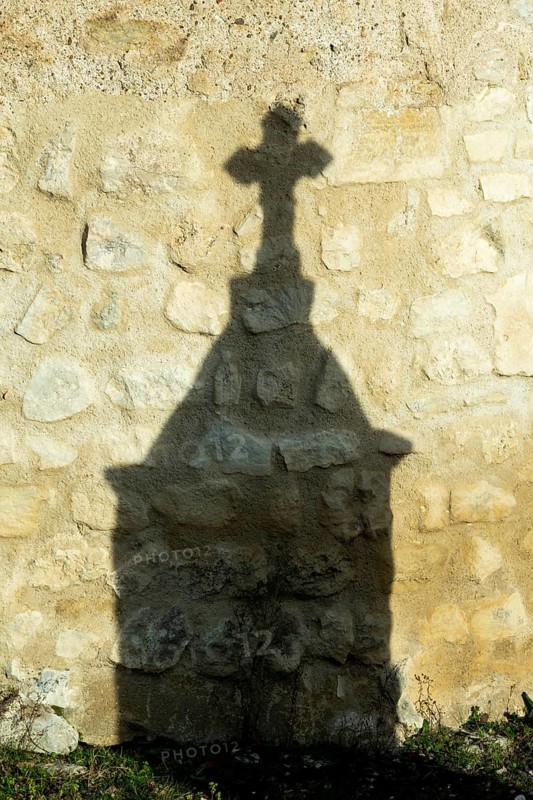 Mazerier village. A shadow of a stone cross appears on an old, weathered stone wall. Sunlight highlights the wall of St. Saturninus Church, Allier department in France. Europe