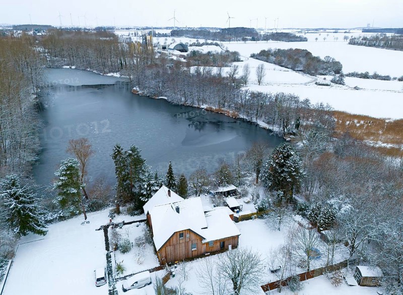Winter landscape with a frozen lake and a snow-covered house on the shore, surrounded by forest, aerial view, Oelsburg, Ilsede municipality, Peine district, Lower Saxony, Germany
