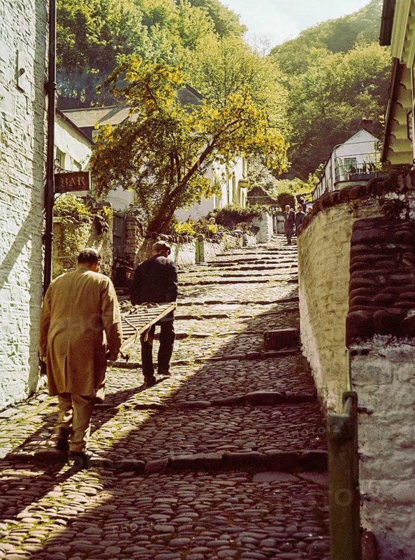 Men carrying wooden sled or sledge used for transport on steep hill of cobbled main village street, Clovelly, north Devon, England, UK 1960s
