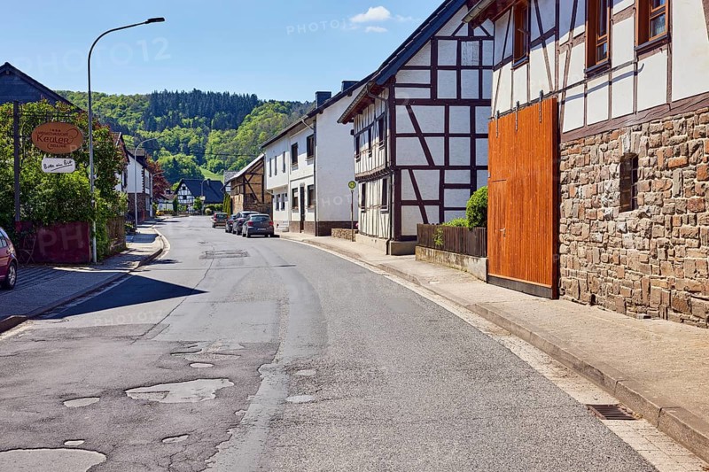 Residential and commercial buildings, historic half-timbered houses with sandstone base, entrance, sidewalk, street, parking strip with cars, hills, hilly landscape, forest, pale blue sky, cumulus clouds, cirrostratus clouds, main street, insul, district of Ahrweiler, Rhineland-Palatinate, Germany