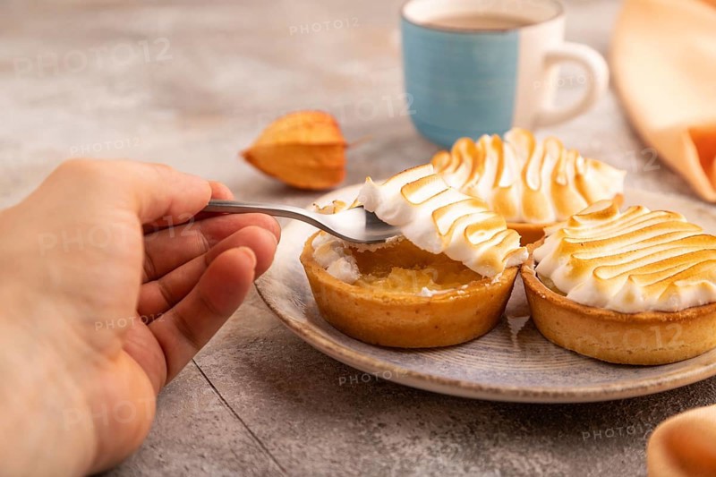 French lemon tart with meringue with hand on brown concrete background, cup of coffee, orange linen textile, side view, close up, selective focus