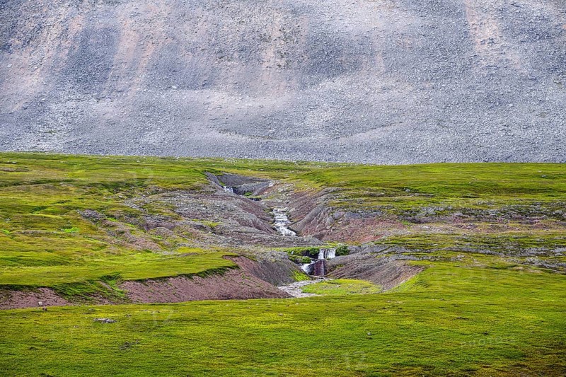 On the slope of a massive granite rock with rugged boulders at Austertanaveien 890 in the foreground, there is a torrent that cuts through green, flat fur beds, green low fell vegetation wide, quiet landscape, Tana, Finnmark, Norway
