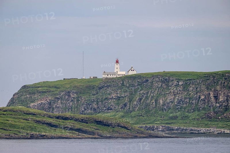 White lighthouse with red tip and antennas rises above steep rocks on the bird island of Hornoya, surrounded by grassy hills under clear skies, Vardø, Finnmark, Norway
