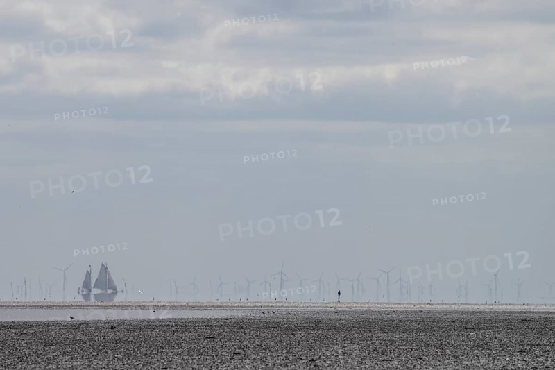 Lonely mudflats scene with wind turbines and sailing ships in the distance, grey landscape with wind turbines and boats in the cloudy sky and lonely mudflat walkers in the haze, Oosterend, Noord-Holland, the Netherlands