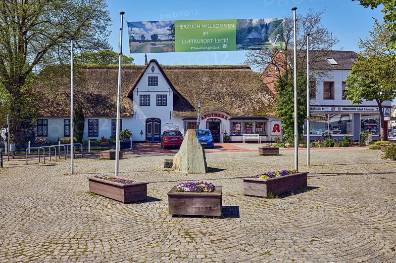 Advertising banners Welcome to the health resort of Leck!, ZumGlückgibtsleck, royally privileged pharmacy, Frisian house, general architecture, flagpoles, flower pots, boulder, trees with spring leaves, square of paving stones, pale blue sky, cirrostratus clouds, main street, market, leak, district of North Frisia, Schleswig-Holstein, Germany