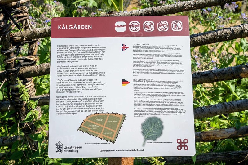 Information sign about Kålgården with texts in various languages and symbols, a picturesque garden with flowers and a rural house in the background, the colorful cottage garden vegetable garden of the famous botanist and taxonomist Carl von Linne with a rustic wooden fence under a blue sky, Linnes Rashult, Rashult, Diö, Kronobergs län, Sweden