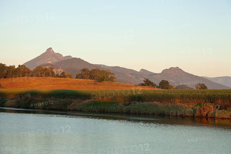 Sunrise light illuminating Mount Warning and surrounding farmland with cultivated cane fields beside a calm river, showing a peaceful agricultural landscape shaped by nature and farming traditions, Murwillumbah, New South Wales, Australia