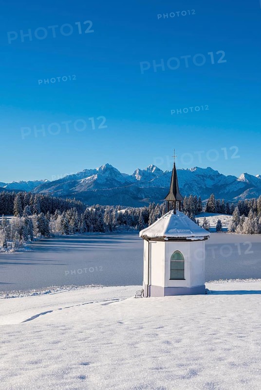 A small chapel against snow-covered mountains and clear blue sky in a winter landscape, Hegratsrieder See, near Füssen, Ostallgäu, Allgäu, Bavaria, Germany