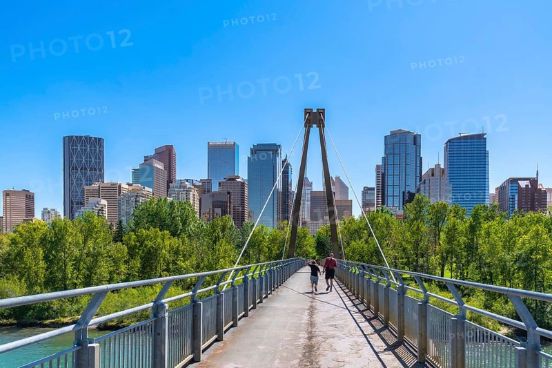 Two tourists are jogging across a pedestrian bridge in calgary, alberta with the downtown skyline visible in the background on a sunny summer day