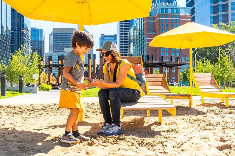 Mother and her son are spending quality time together at an urban beach in calgary, sheltered by yellow parasols, with the city skyline in the background