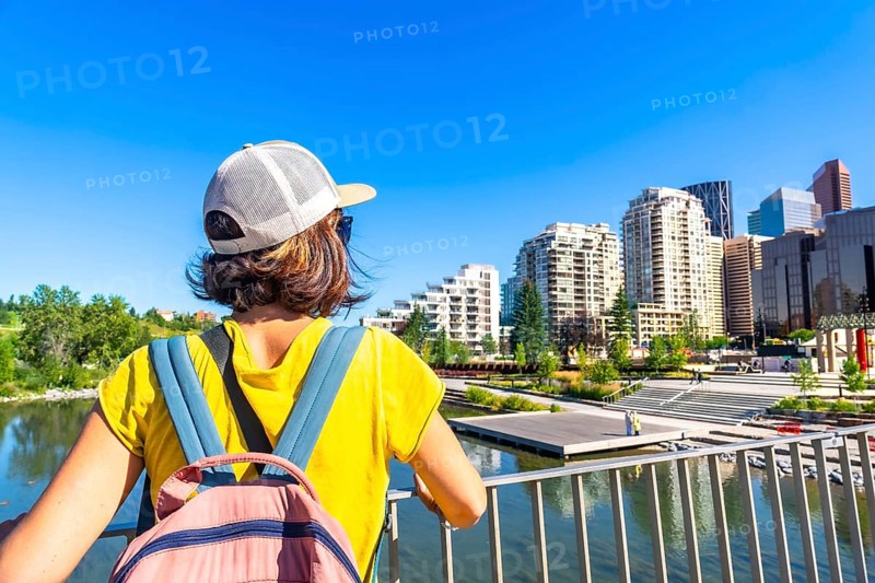Tourist with a backpack is enjoying the cityscape of calgary, alberta, from a bridge, taking in the modern architecture and the bow river on a sunny day