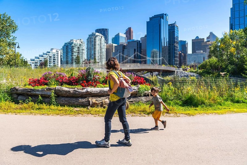 Mother with backpack walking with her running son in calgary's east village riverwalk, enjoying a sunny summer day with the city skyline in the background