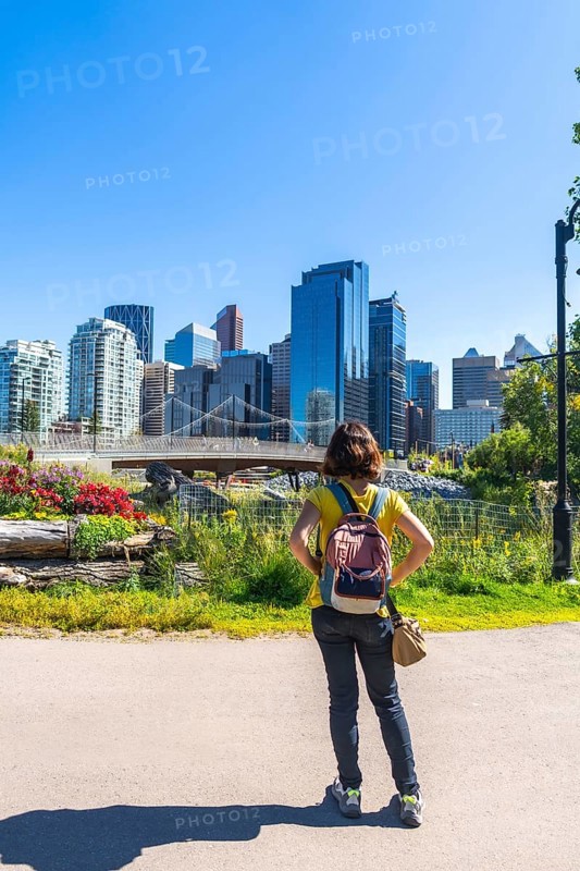 Tourist with a backpack stands in a colorful park, gazing at the modern cityscape of calgary, alberta, under a clear blue sky, enjoying the urban beauty and vibrant atmosphere