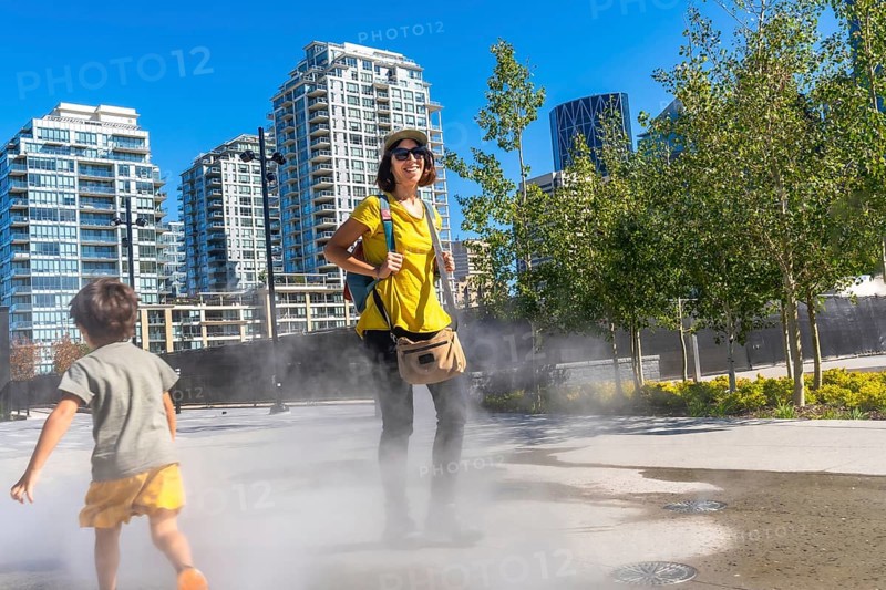 Tourist in a yellow shirt and backpack smiling while child runs through a refreshing misting system on a hot summer day in calgary, alberta, with modern buildings behind