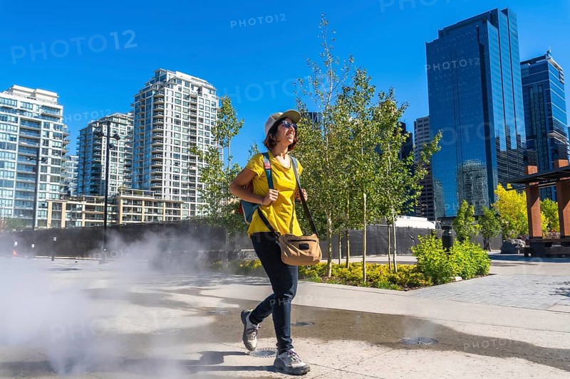 Female tourist walking through a refreshing misting station in calgary's east village, enjoying a sunny summer day while surrounded by urban skyscrapers and vibrant greenery