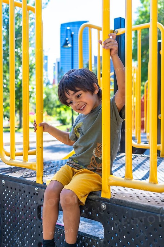 Young boy with a joyful expression sits on a playground structure, holding onto yellow bars and smiling, enjoying a sunny day in a city park in calgary, canada