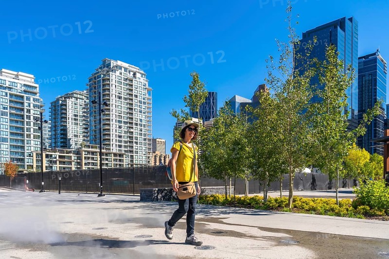 Tourist wearing a yellow t shirt and a hat is enjoying a walk in calgary's east village neighborhood on a sunny day, with modern residential buildings and skyscrapers in the background