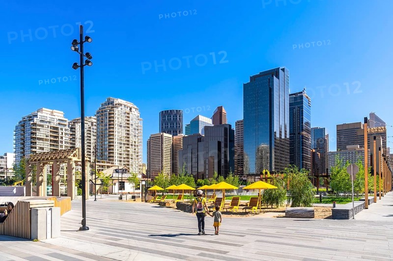 Modern skyscrapers rise above a newly developed urban park in calgary's east village, where a parent and child are enjoying a stroll on a sunny summer day